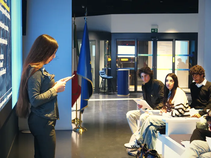 Una partecipante pronuncia il suo discorso accanto alle bandiere dell’Unione europea e dell’Italia, mentre il gruppo ascolta.